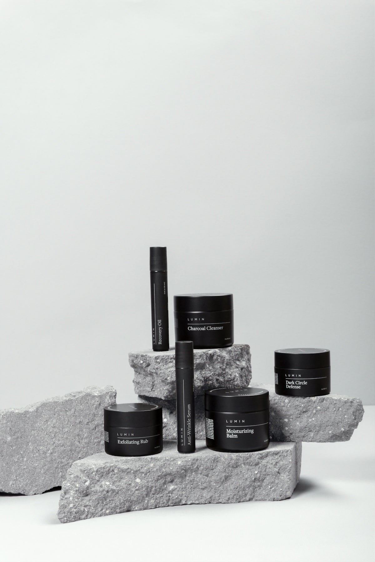 Six black skincare containers arranged on stacked grey stone blocks against a light background