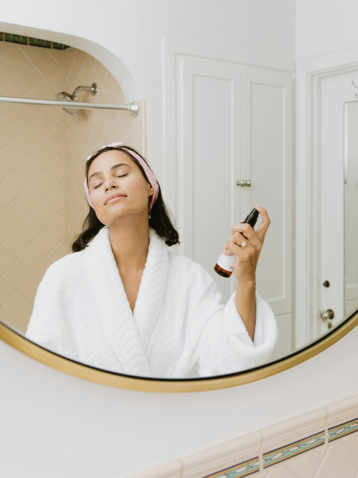 A woman in a white robe and headband spraying facial mist in a bathroom