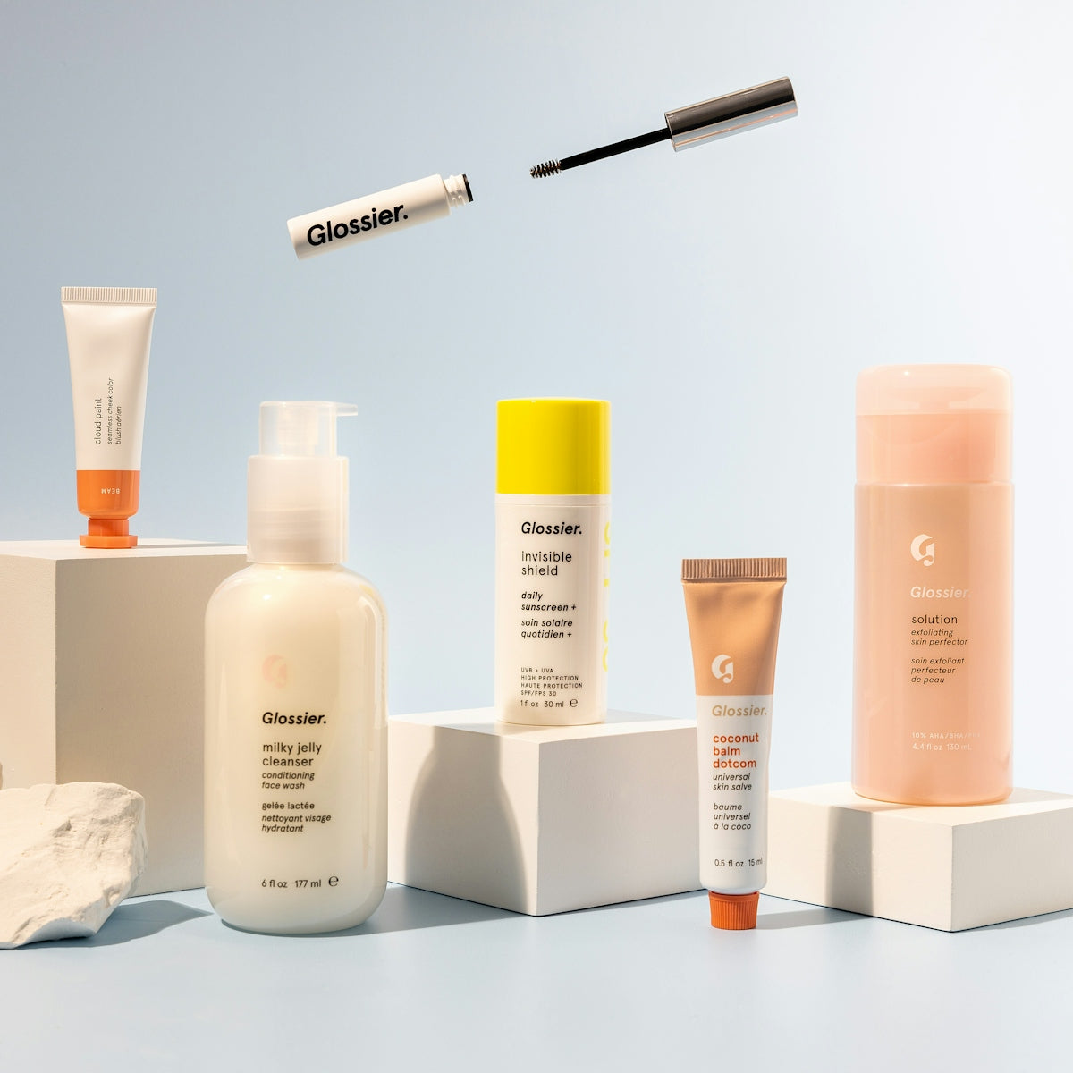 Assorted skincare products and a mascara wand arranged on white pedestals against blue